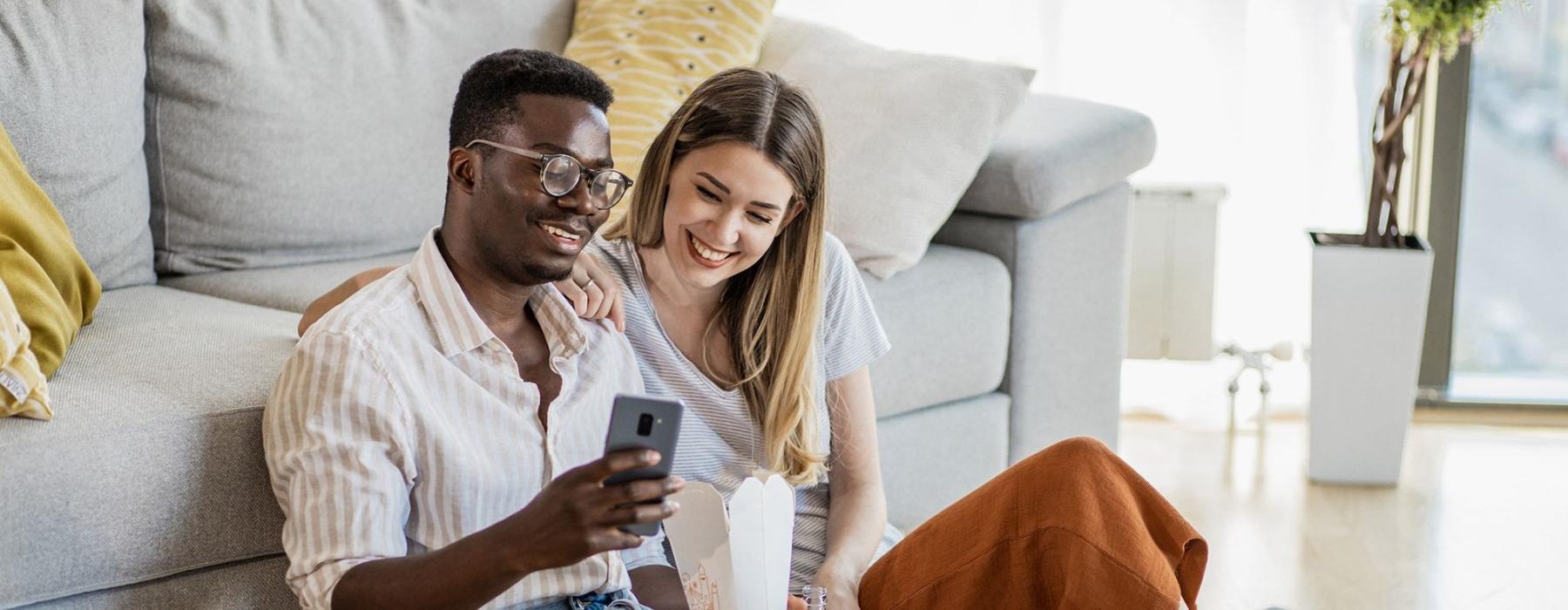 a man and woman with take out, sit against a couch on their living room floor and watch at their cell phone