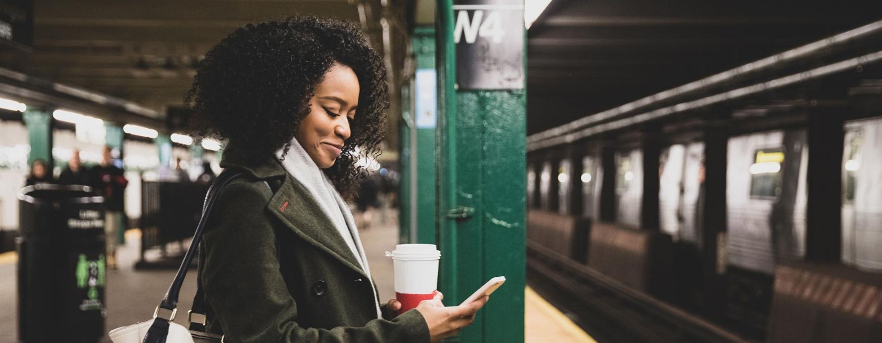 woman with a cup of coffee texts on subway station platform